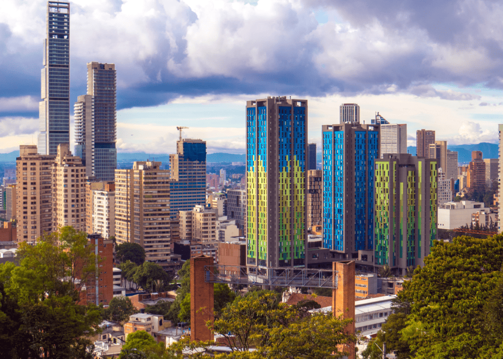 The modern skyline of Bogotá Colombia featuring colorful glass towers and historic buildings set against a dramatic cloudy sky with the Andes in the background.