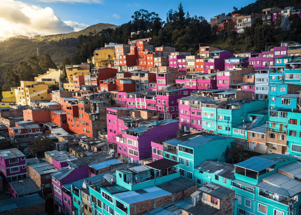An aerial view of a densely packed hillside neighborhood in Bogotá Colombia with buildings painted in vivid shades of pink teal orange and purple at golden hour.