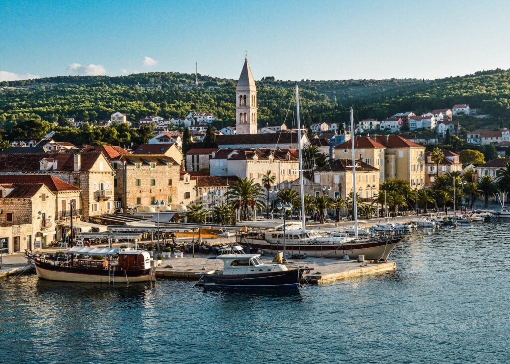Stone waterfront of Brač Island in Croatia at golden hour with sailboats moored at the dock and a church bell tower rising above the palm-lined harbor