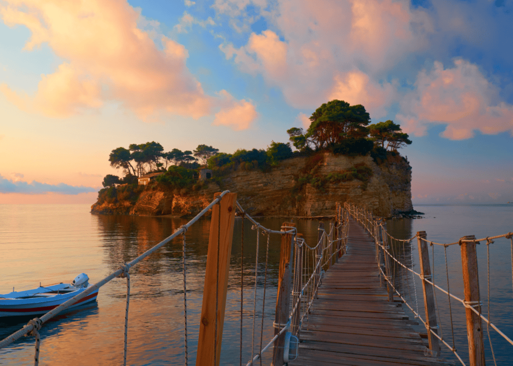 A wooden rope bridge stretches across calm water toward Cameo Island in Zakynthos Greece at sunset with pine trees crowning the rocky islet and pink and golden clouds reflected in the still sea.