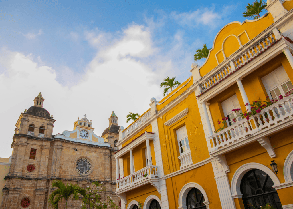 A bright yellow colonial building with white balconies and flowering bougainvillea stands beside a stone church in a sun-filled plaza in Cartagena Colombia.