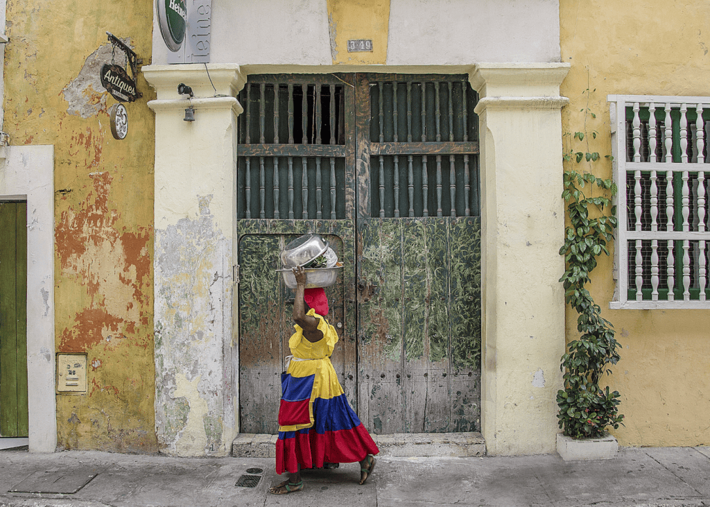 A palenquera woman in a traditional red yellow and blue ruffled dress carries a bowl on her head while walking past a weathered colonial doorway in Cartagena Colombia.