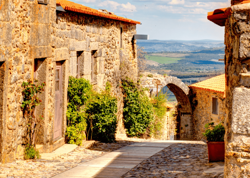 A narrow cobblestone lane in the historic village of Castelo Rodrigo Portugal with warm golden granite houses on either side, a stone arch framing climbing green plants, and a sweeping view of the Côa Valley countryside stretching into the distance.