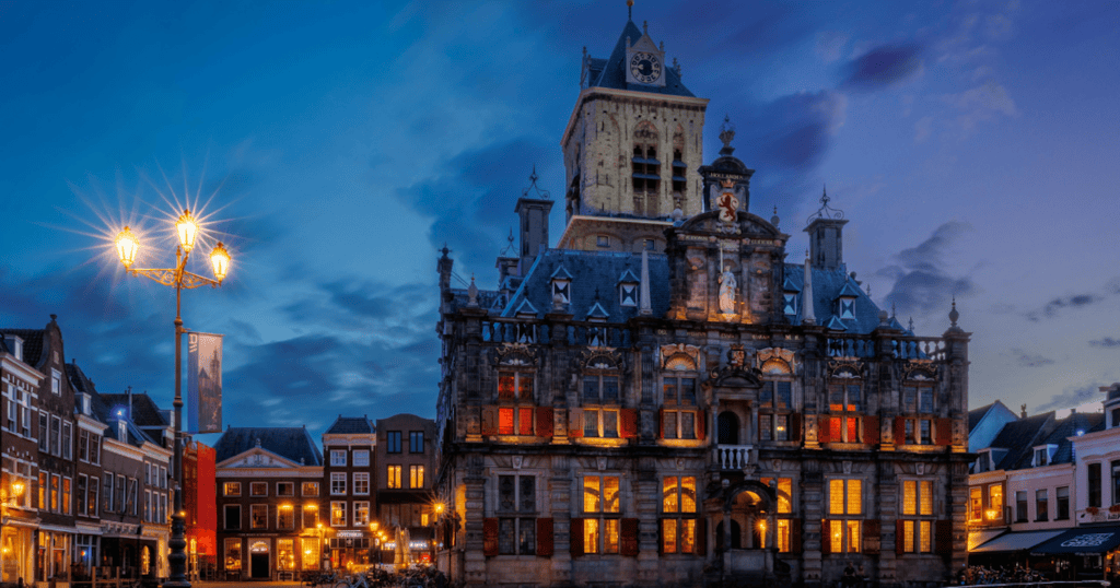 The ornate Renaissance facade of Delft's town hall glows with warm golden light at dusk, illuminating the Markt square with its cobblestones and surrounding historic buildings under a deep blue evening sky.