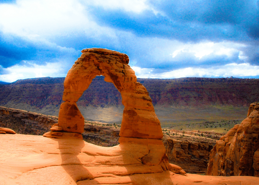 reestanding Delicate Arch rising from red sandstone with canyon views and dramatic cloudy sky in Arches National Park Utah.