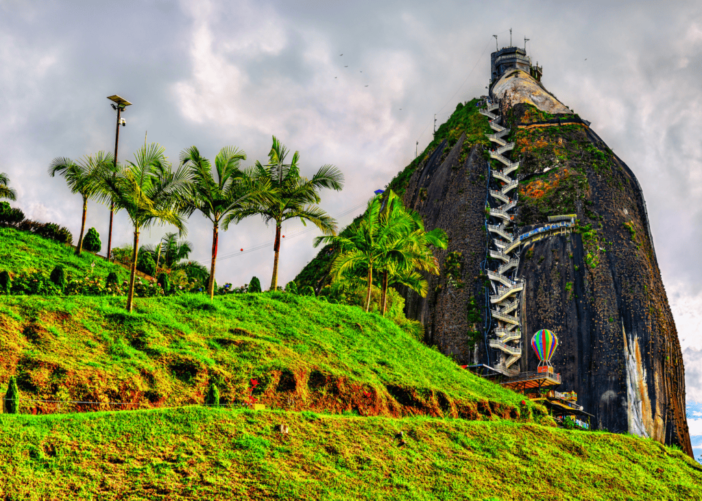 The massive rock of La Piedra del Peñol in Guatapé Colombia with a zigzagging staircase built into its side rising above green hillsides and palm trees.
