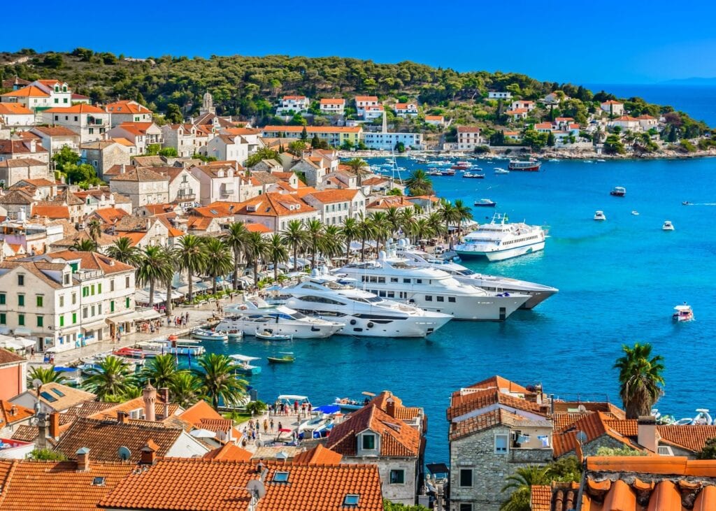 Aerial view of Hvar Town harbor in Croatia with luxury yachts moored along the palm-lined waterfront and terracotta rooftops spreading up the hillside above the turquoise Adriatic
