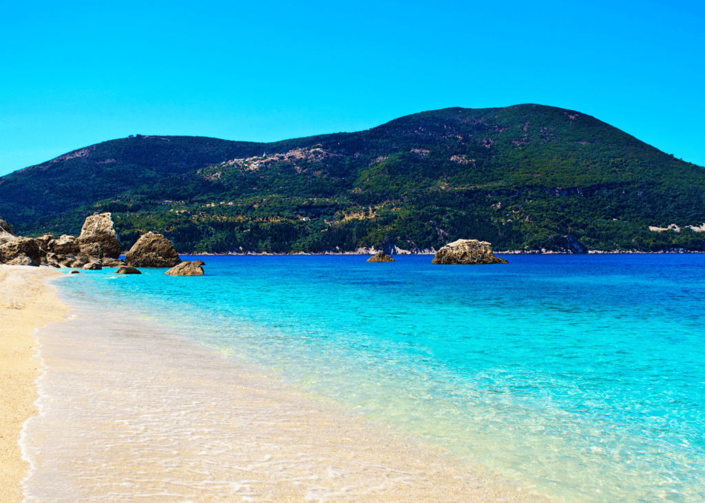 A wide sandy beach on a Greek Ionian island stretches toward turquoise water with scattered rocks and a lush green mountain rising along the coastline under a vivid blue sky.