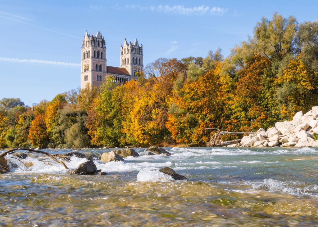 Isar River in Munich Germany rushing over rocks in autumn with golden orange foliage and twin towers of a Romanesque church rising above the trees.