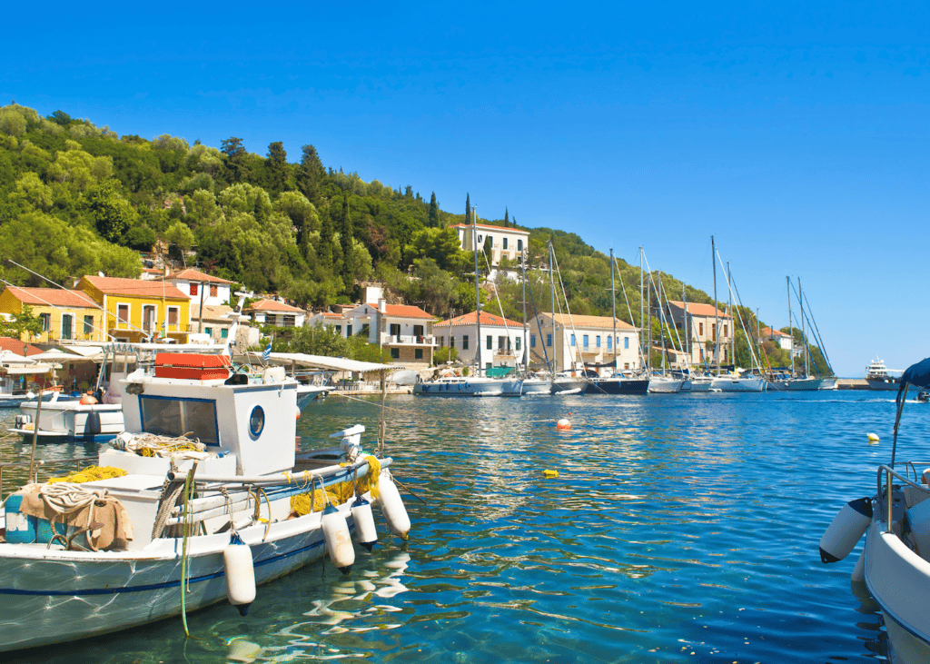 Fishing boats and sailboats bob in the calm clear harbor of a small village on Ithaca Greece with colorful waterfront buildings and densely forested hills rising behind them.