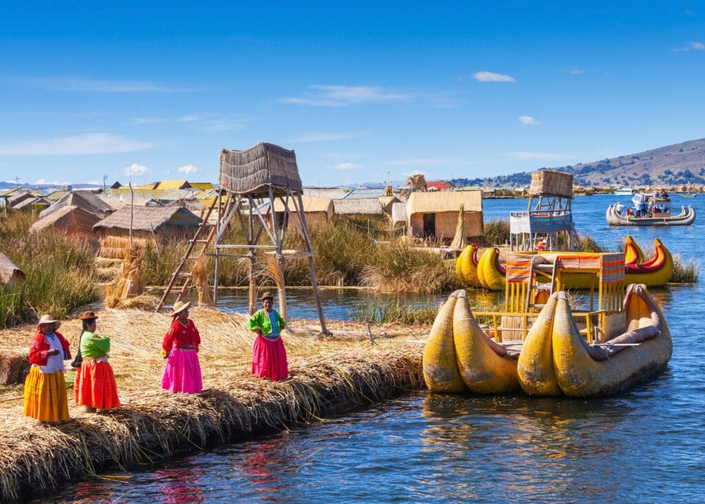 Uros women in colorful traditional skirts standing on a floating totora reed island on Lake Titicaca Peru with reed boats and thatched structures behind them