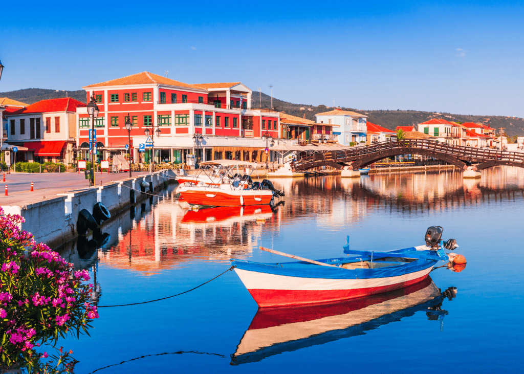 A colorful red and white fishing boat reflects in the calm harbor water of Lefkada Town Greece with vibrant buildings a wooden bridge and pink oleander flowers lining the waterfront promenade.
