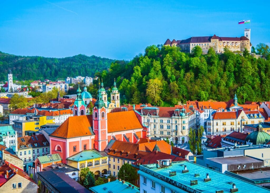 Panoramic view of Ljubljana Slovenia showing colorful orange rooftops, the pink Franciscan Church, and Ljubljana Castle sitting on a green forested hill under a bright blue sky in spring.