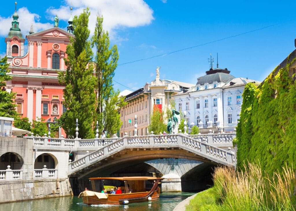 A wooden boat passing under an ornate stone bridge on the Ljubljanica River in Ljubljana, Slovenia, with the pink Franciscan Church and colorful historic buildings in the background on a sunny summer day.