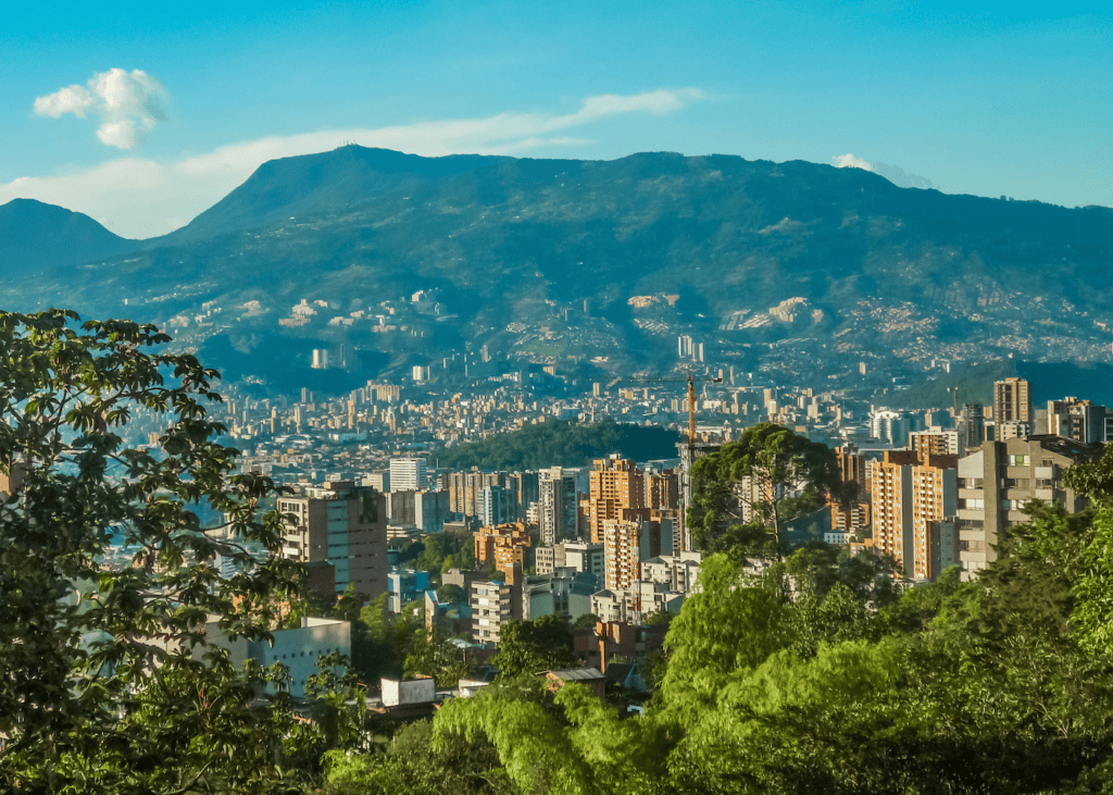 A wide view of Medellín Colombia nestled in a lush green valley in the Andes mountains with city buildings and tropical trees in the foreground.