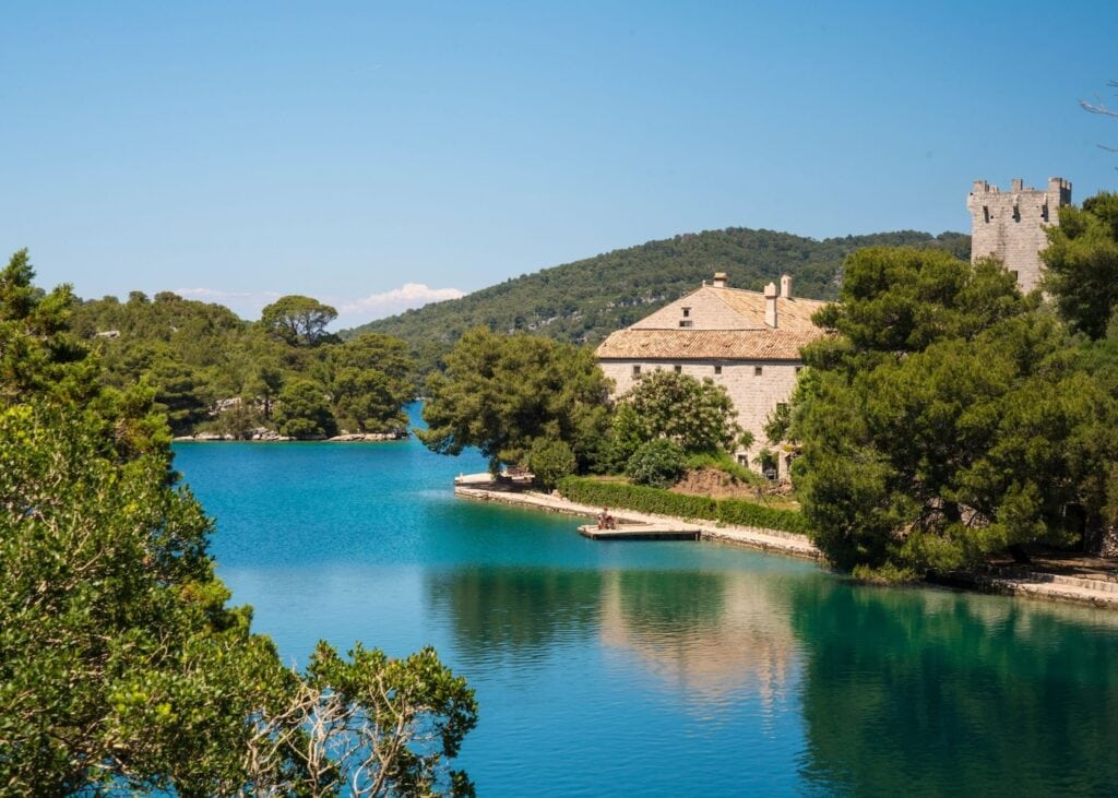 Vivid blue saltwater lake at Mljet National Park in Croatia with a historic stone monastery and medieval tower reflected in the calm water surrounded by dense pine forest