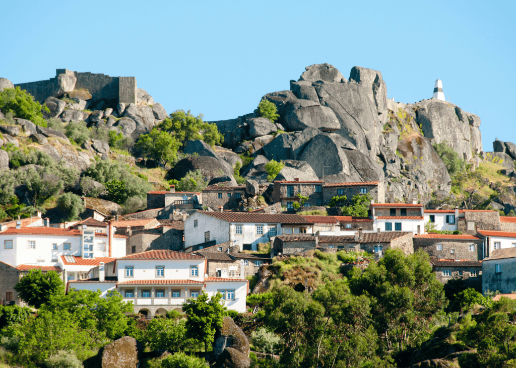 The historic village of Monsanto Portugal built among massive granite boulders on a steep hillside with a medieval castle ruin visible at the top against a bright blue sky.