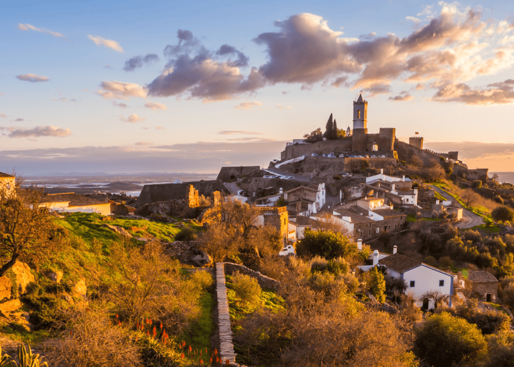 The medieval hilltop village of Monsaraz Portugal bathed in golden sunset light with whitewashed houses, a castle tower, and the vast Alentejo landscape stretching out beneath a dramatic cloud filled sky.