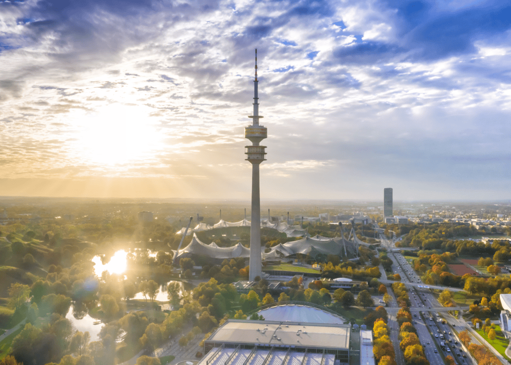 Aerial view of Olympiapark in Munich Germany at sunset with the Olympic Tower and iconic tent roof structures surrounded by autumn trees.
