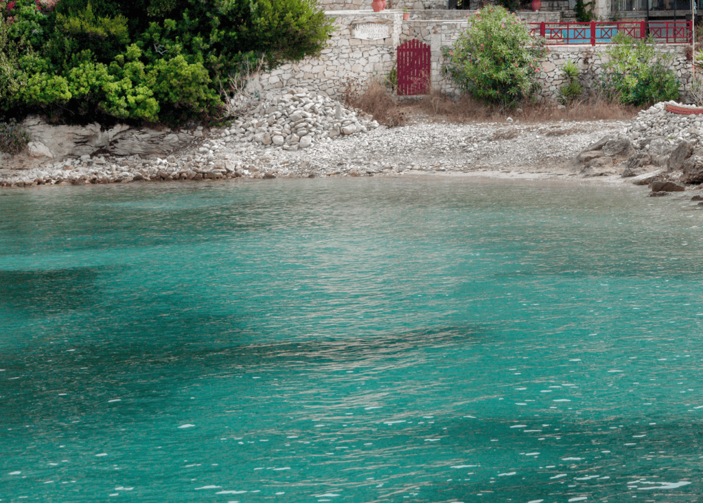 Crystal clear turquoise water laps a quiet pebble cove in the Ionian Islands of Greece with lush greenery and a traditional stone building along the shore.