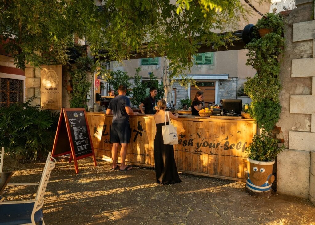 Warm golden light falls on an outdoor food and coffee bar in a lush courtyard in a Croatian old town with stone walls and green vines framing the scene