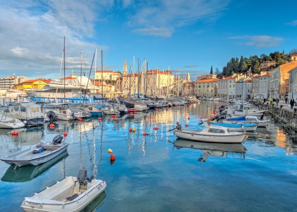 The harbor of Piran Slovenia on a clear winter day with small boats and sailboats reflected in calm blue water and the historic old town and church bell tower visible in the background.