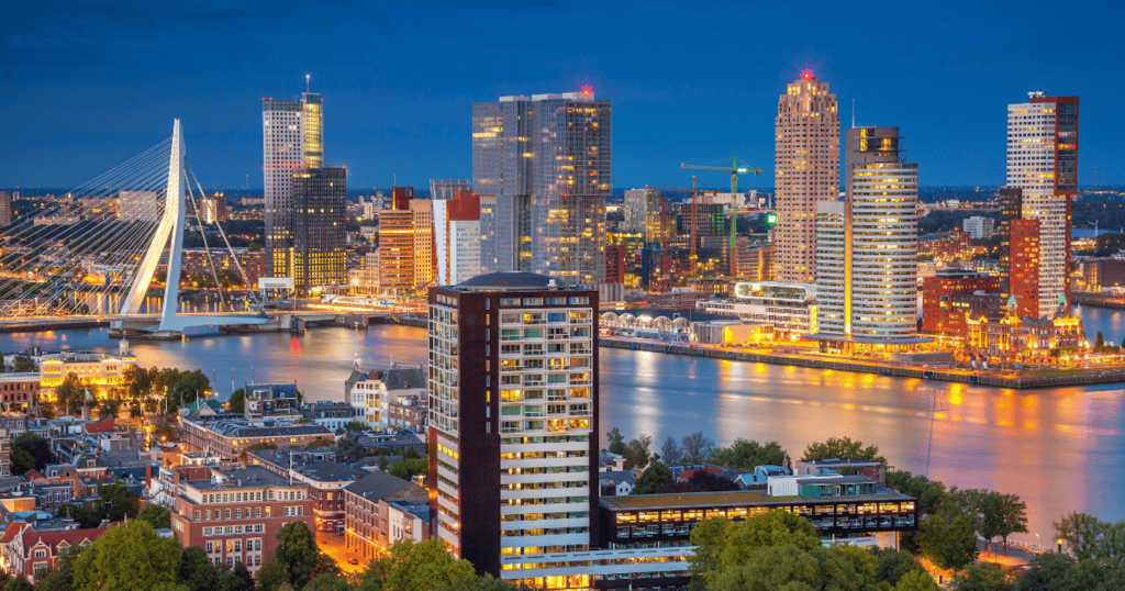 The dramatic Rotterdam skyline at night reflected in the Maas River with the white cable-stayed Erasmus Bridge on the left and illuminated modern skyscrapers lining the waterfront under a deep blue sky.