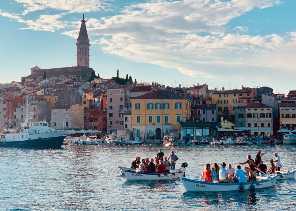 mall wooden boats carrying passengers across the harbor in Rovinj Croatia with colorful buildings and the tall church tower of St Euphemia rising above the town at sunset