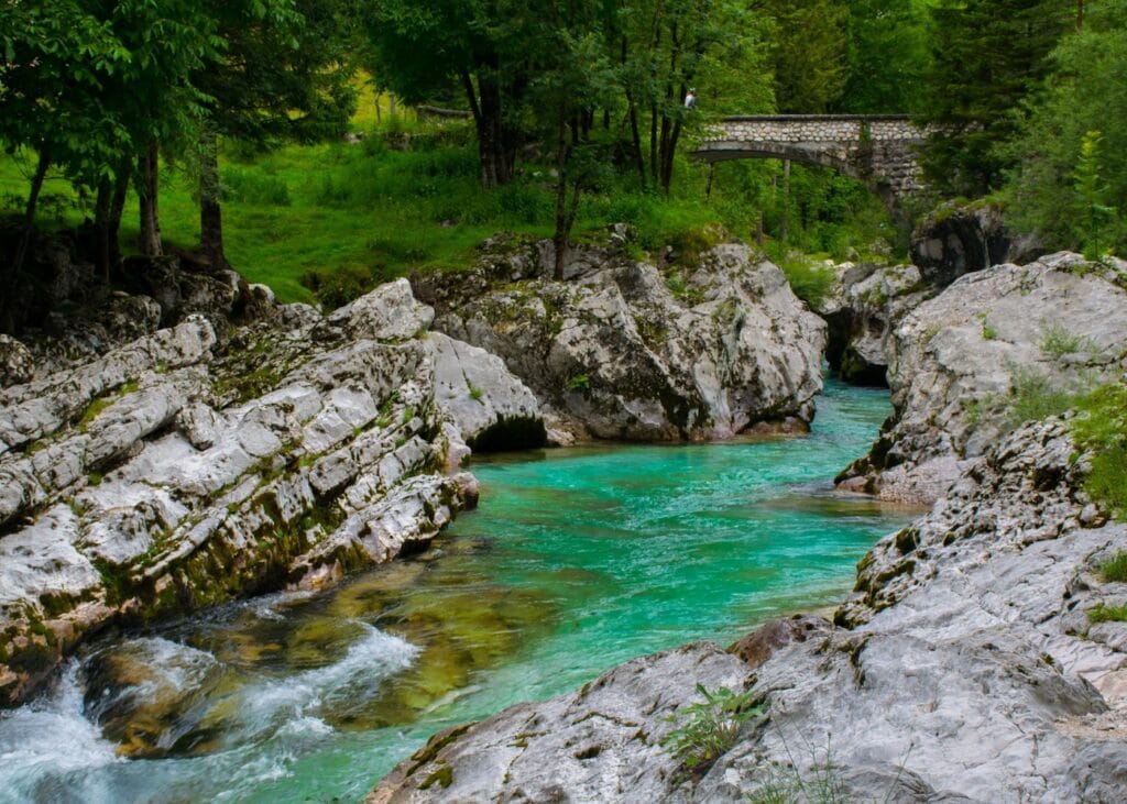The turquoise Soča River rushing between large white limestone rocks in a lush green forest valley in Slovenia, with a stone bridge visible in the background.