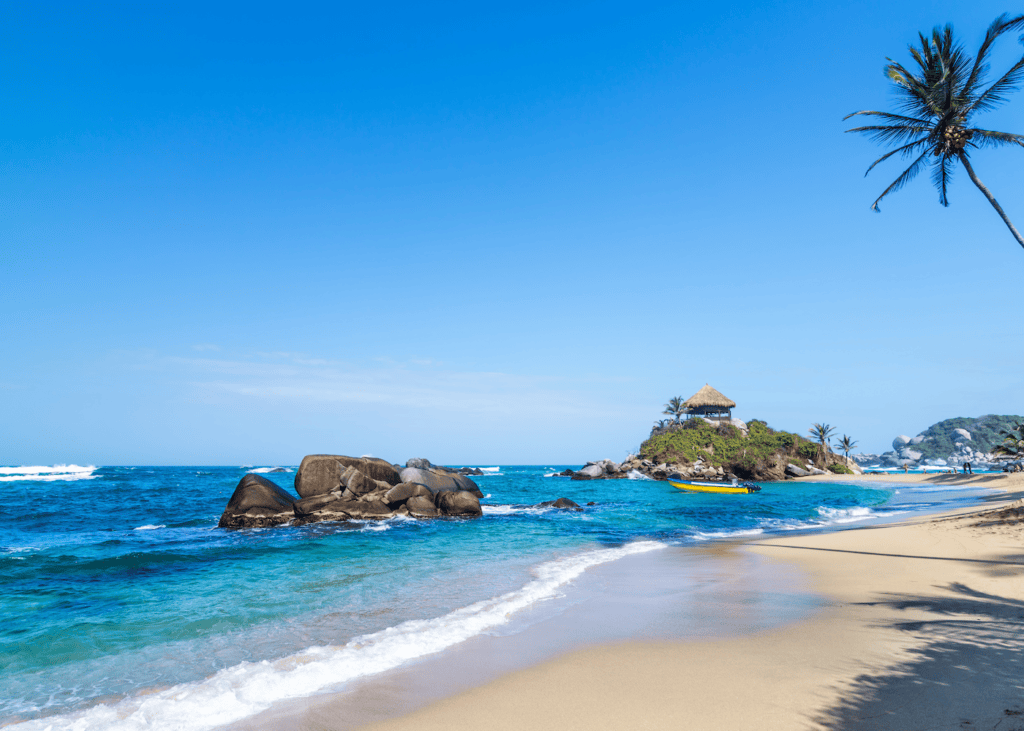 A pristine beach in Tayrona National Park Colombia with turquoise water smooth boulders a leaning palm tree and a thatched hut perched on a rocky point under a clear blue sky.