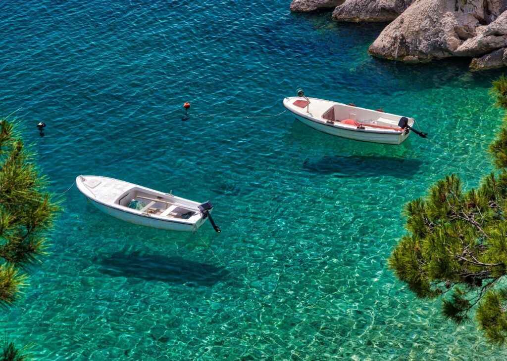 Two small white boats floating in crystal clear turquoise Adriatic water off the Croatian coast with rocky shoreline and pine trees visible above
