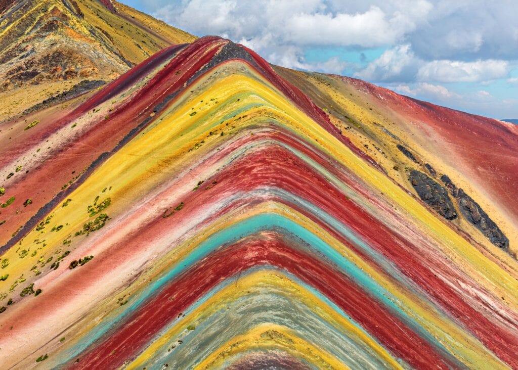 Close up of Vinicunca Rainbow Mountain Peru showing vivid mineral stripes of red yellow green and teal across the mountain ridge under a cloudy sky