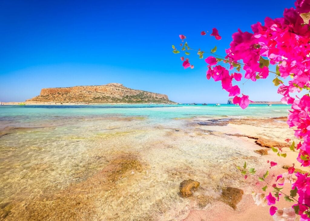 Stunning shallow turquoise lagoon at Balos, Crete with pink bougainvillea framing the view toward a rocky island