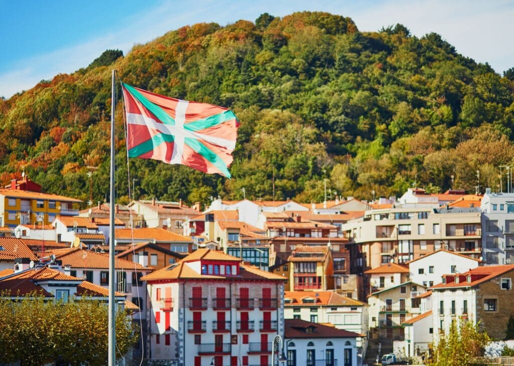 The red, green and white Basque flag flying over a hillside village in Basque Country Spain with autumn foliage covering the green hill behind the rooftops.
