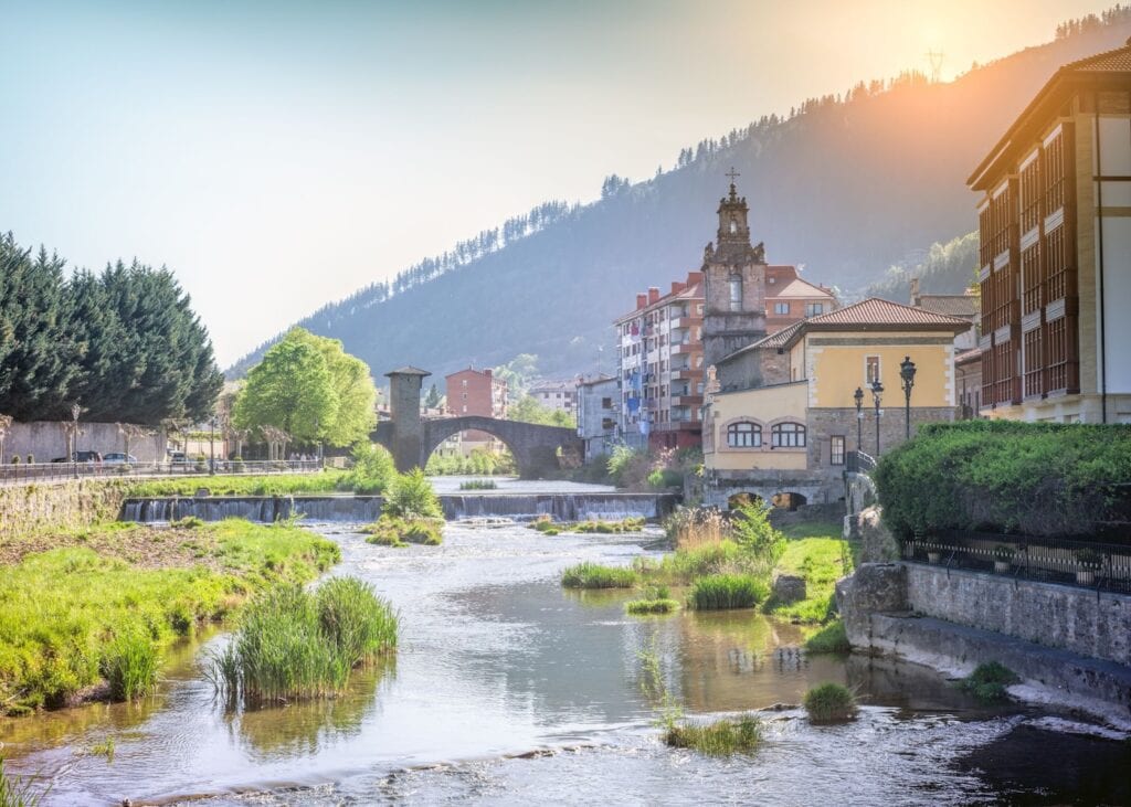 A stone arched bridge spanning a gentle river through a Basque Country village in northern Spain with a church steeple and forested mountains glowing in golden sunset light.