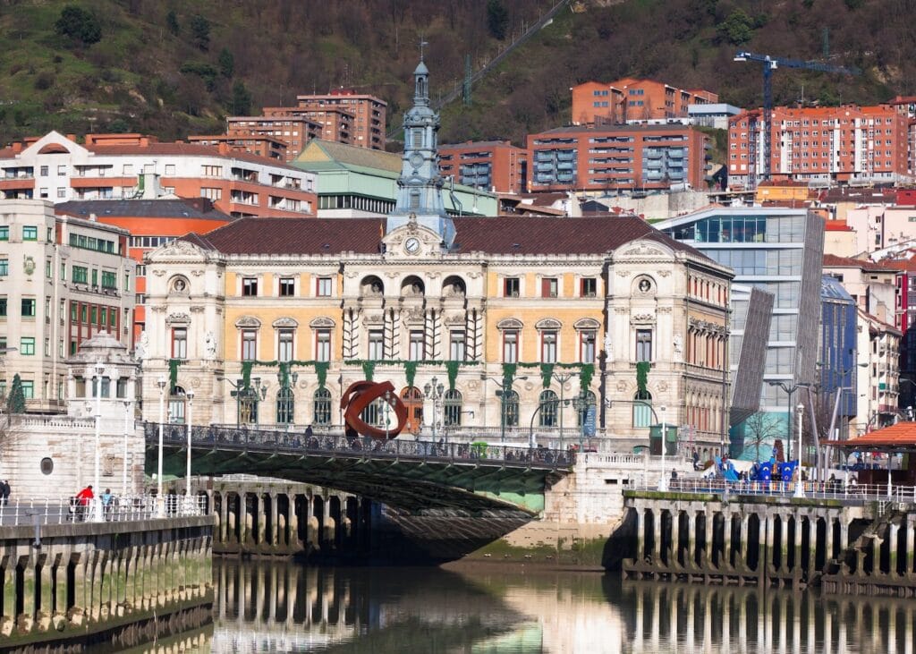 The ornate beaux arts facade of Bilbao city hall reflected in the Nervión River in Bilbao Spain with a modern footbridge visible to the right.