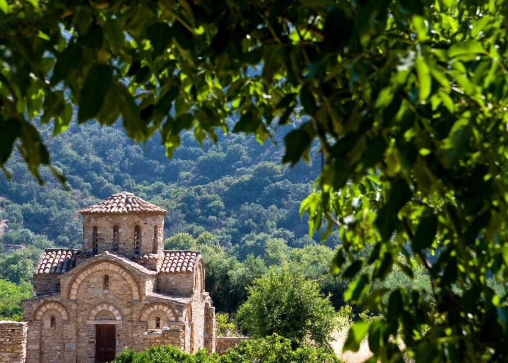 Ancient Byzantine stone church nestled among lush green hills in the Cretan countryside framed by tree branches