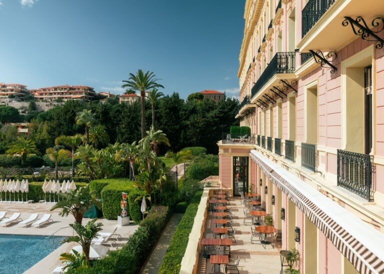 Exterior view of Royal Riviera hotel's pink Belle Epoque facade with pool terrace, white sun loungers, striped awnings, and lush tropical gardens