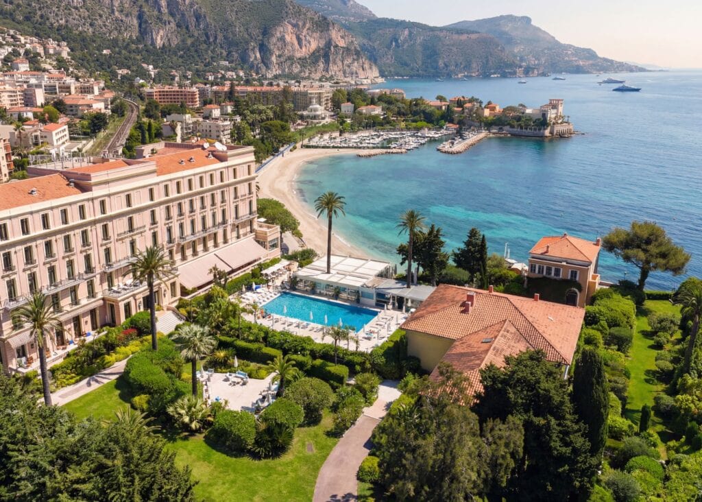 Aerial view of Royal Riviera hotel in Saint-Jean-Cap-Ferrat with outdoor pool, palm-lined gardens, private beach, and turquoise Mediterranean bay