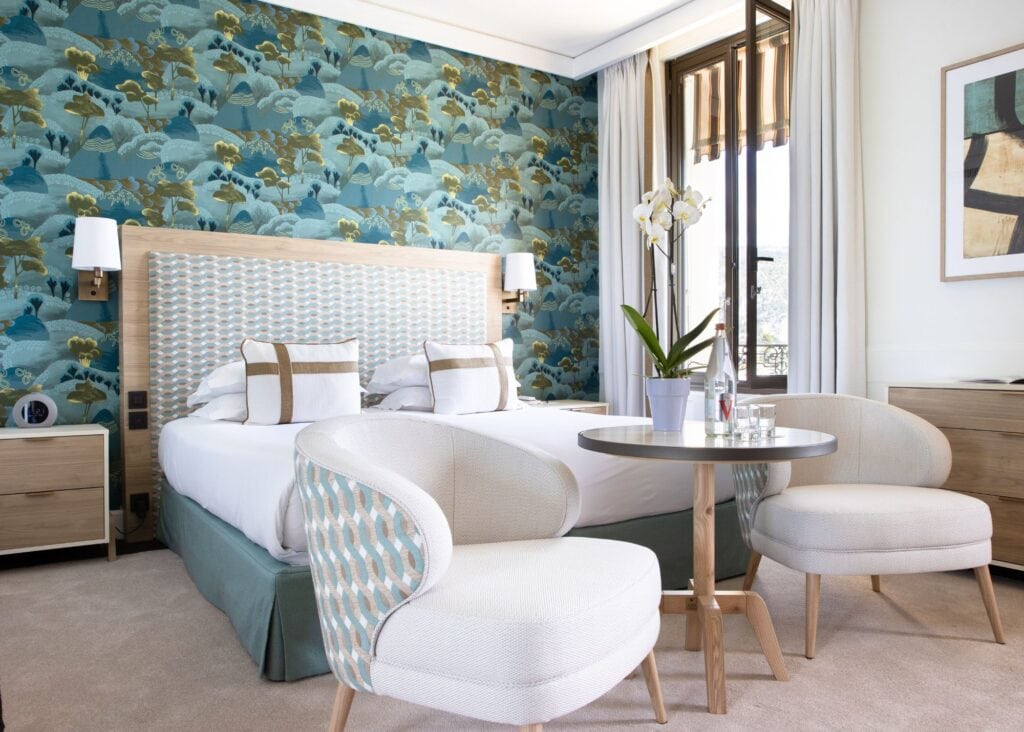 Deluxe guest room at Royal Riviera hotel with bold blue botanical wallpaper, upholstered headboard, curved cream armchairs, and French doors with garden light