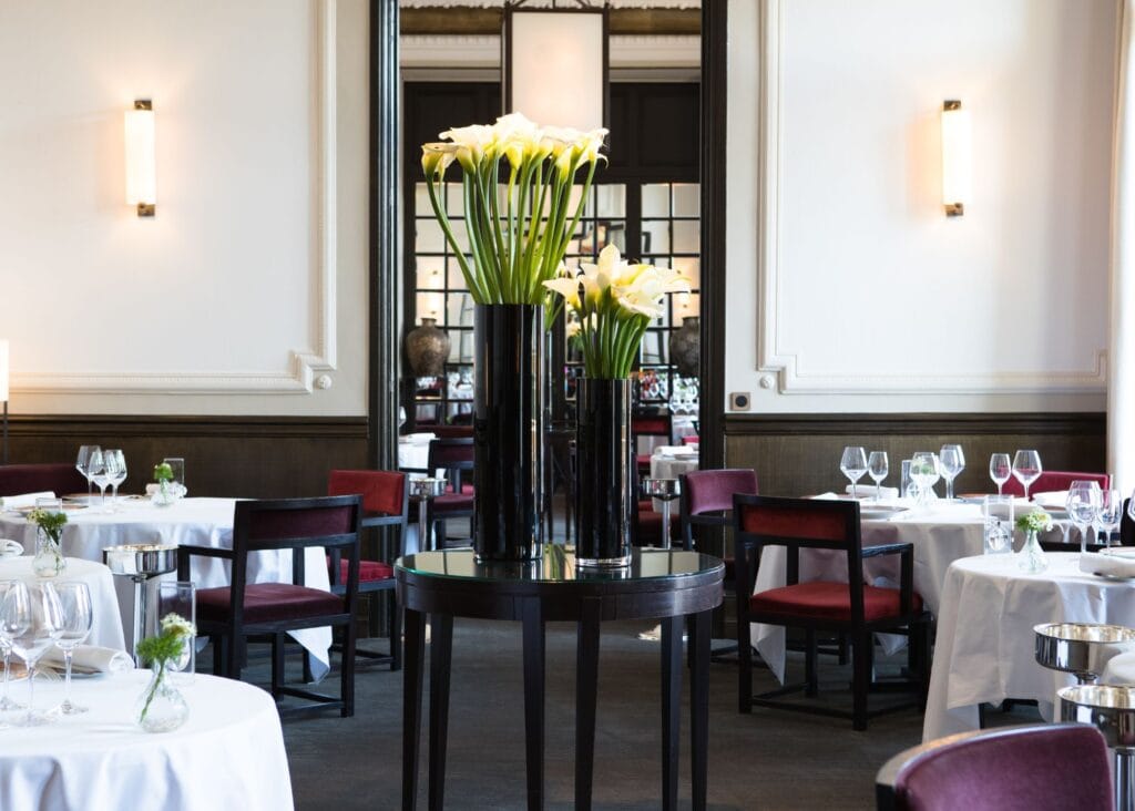 Formal dining room at Royal Riviera hotel with white-clothed round tables, burgundy upholstered chairs, tall calla lily arrangement, and mirrored wall panels