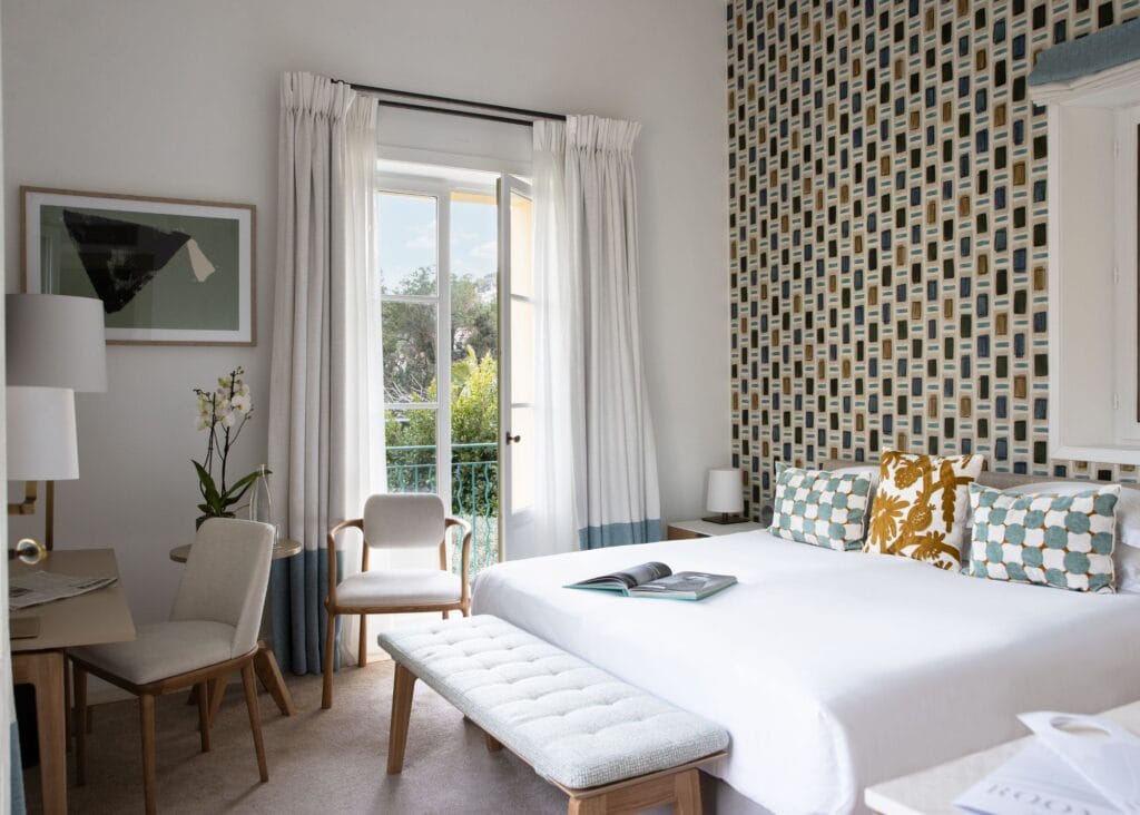 Bright guest room at Royal Riviera hotel with patterned accent wall, white bedding, teal and gold throw pillows, and French doors opening to a garden view