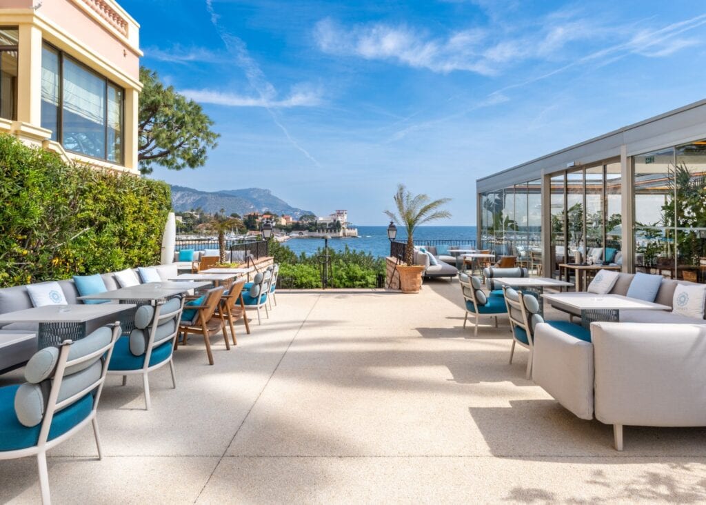Spacious outdoor lounge terrace at Royal Riviera hotel with blue and white upholstered seating, teak chairs, and unobstructed views over the bay toward the sea