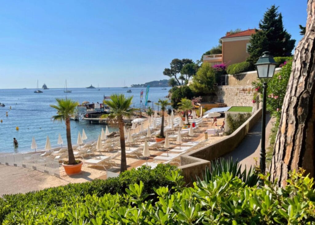 View from the garden path at Royal Riviera hotel looking down to the private beach, white loungers, palm trees, and sailboats anchored in the bay beyond