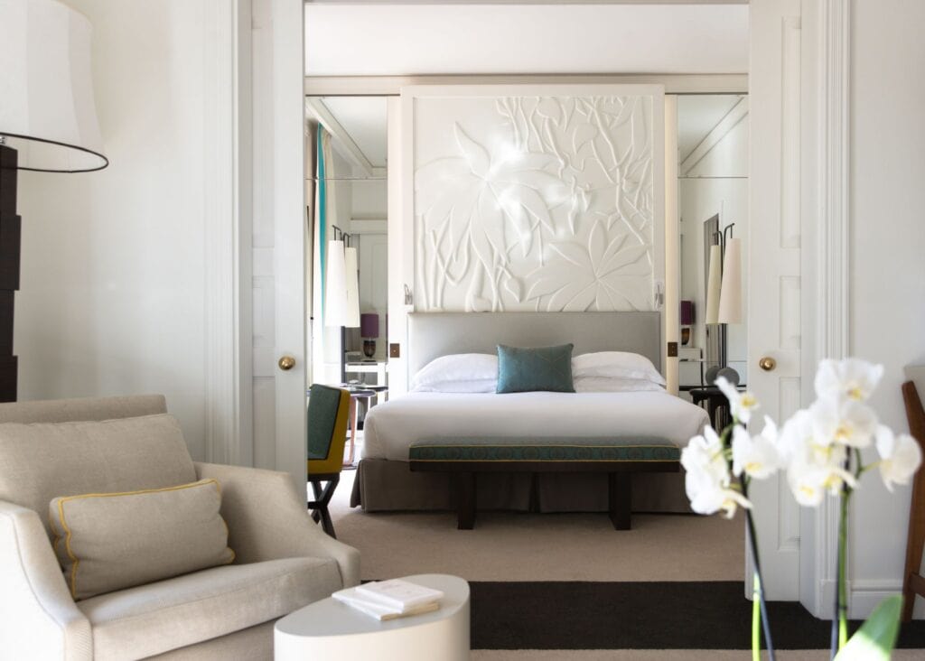 Suite at Royal Riviera hotel featuring a dramatic white sculptural floral headboard, mirrored walls, white orchids, and a separate sitting area with cream armchairs