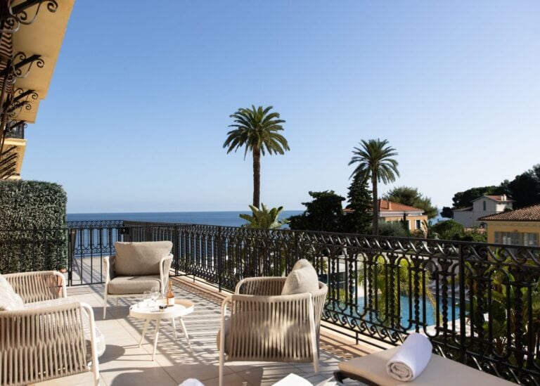 Private suite terrace at Royal Riviera hotel with woven lounge chairs, champagne on ice, pool below, and open Mediterranean Sea view framed by palm trees