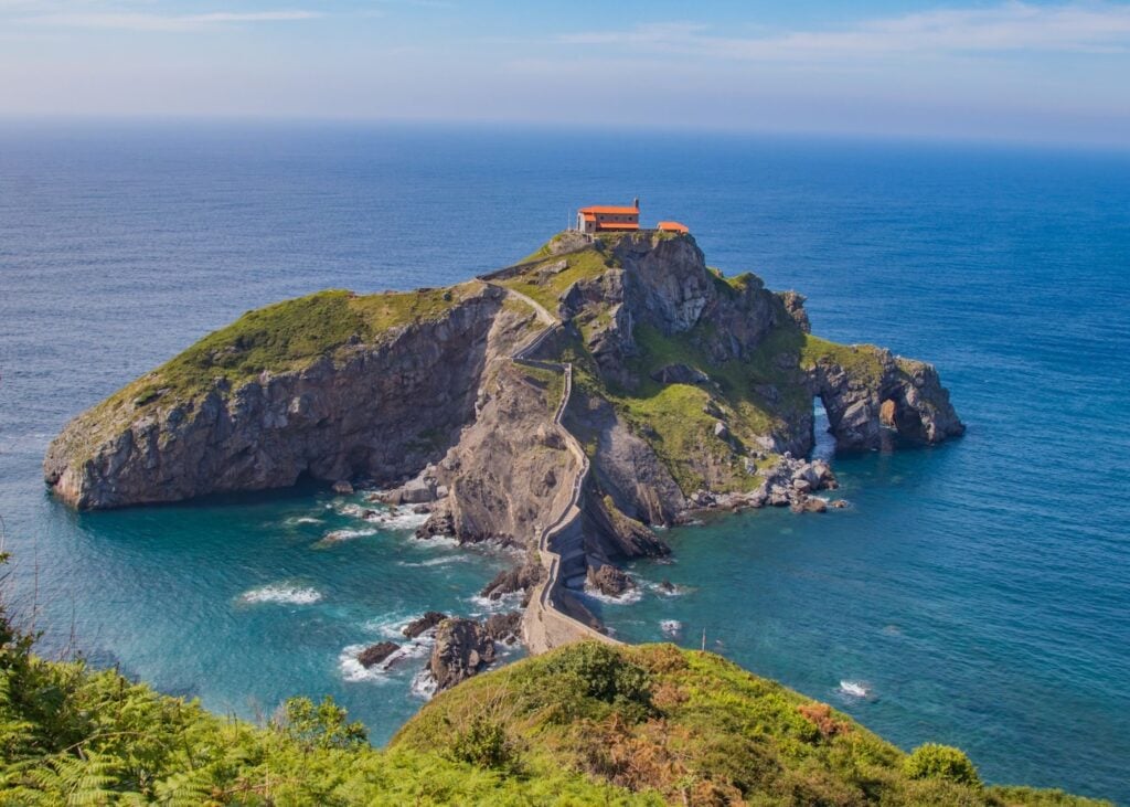 The dramatic rocky island of San Juan de Gaztelugatxe off the Basque Country coast in Spain with a narrow stone stairway path leading up to a small church at the top surrounded by deep blue Atlantic water.