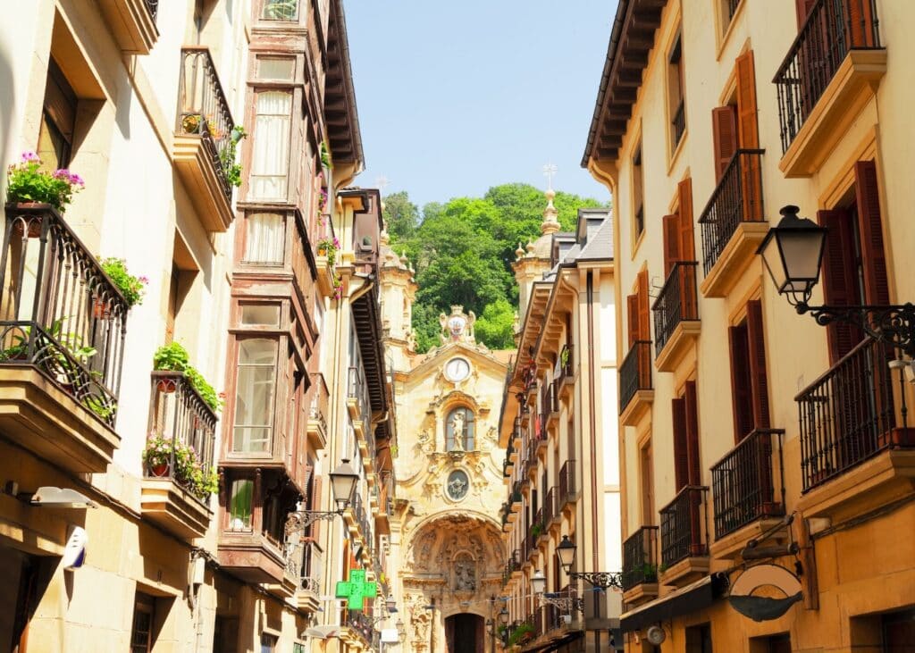 A narrow pedestrian street in the Old Town of San Sebastián Spain lined with ornate balconies and flower boxes with a baroque church facade visible at the end of the lane.