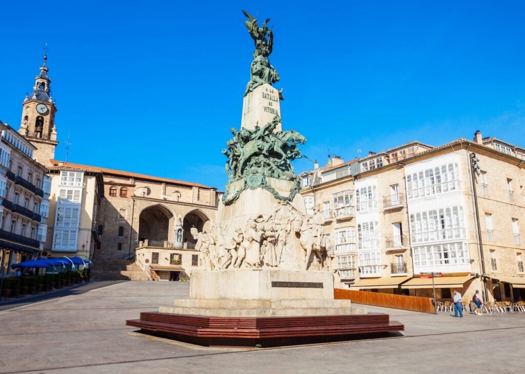 The ornate bronze Battle of Vitoria monument standing in the main plaza of Vitoria-Gasteiz Spain with the medieval church of San Miguel and stone buildings surrounding the open square under a deep blue sky.