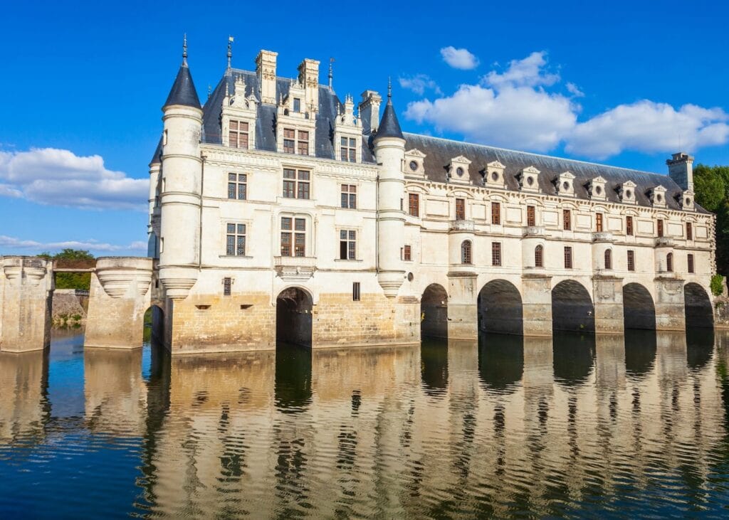 Chateau de Chenonceau spanning the Cher River in the Loire Valley France with its arched stone base reflected in the still water under a bright blue sky